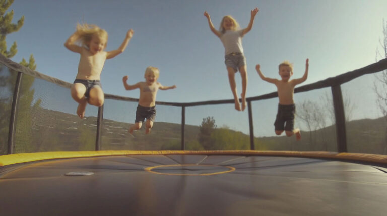 Four young children jumping together on an outdoor trampoline with a safety net and scenic hills in the background